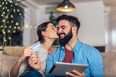 A couple sits on a couch and smile while using a credit card to buy stuff on their iPad.