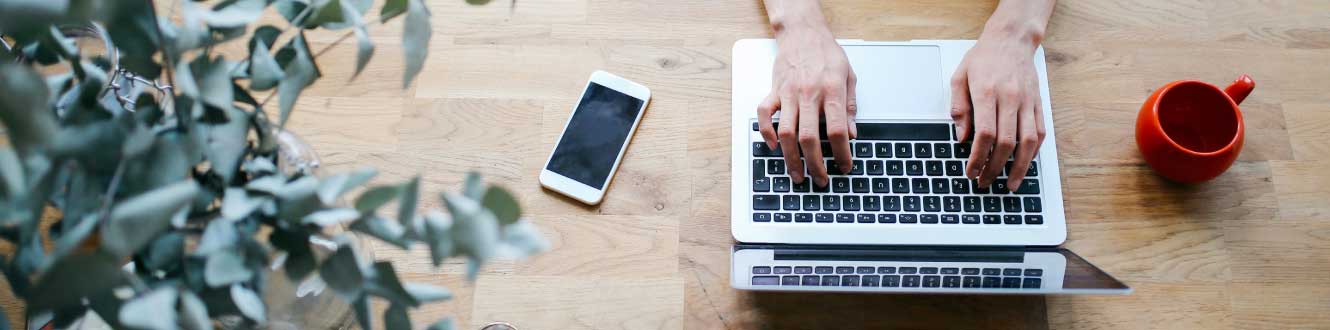 A top down view of a woman typing on her laptop with a cup of coffee and mobile phone also in view.