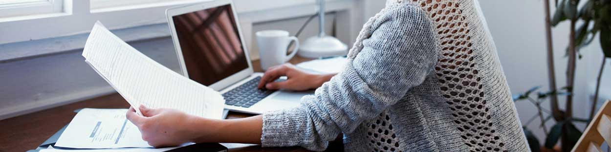 A woman looking at paperwork while typing on a laptop.