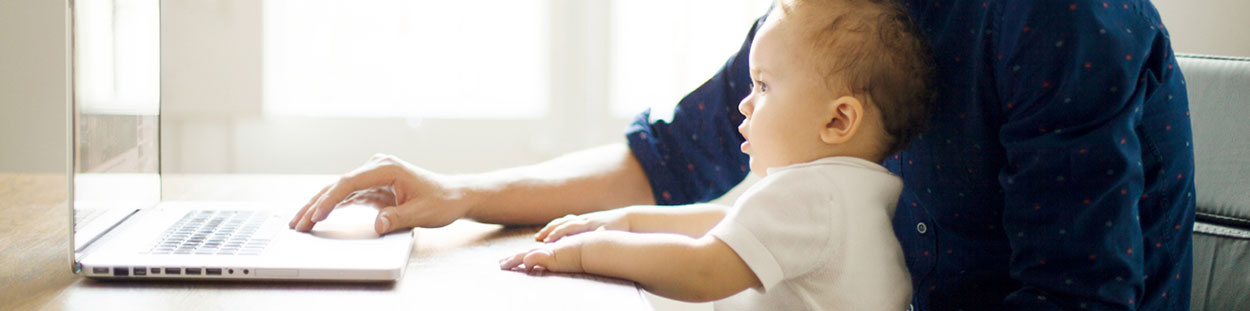 A father with his baby on his lap while he is typing at his desk.