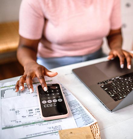 Woman using a calculator on her phone sitting at her desk in front of a laptop.